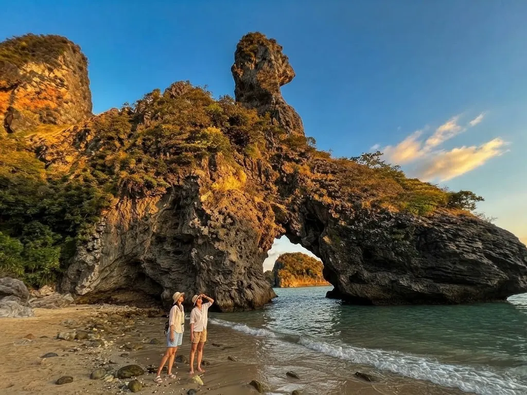 Two travellers on the beach under a natural rock arch on Chicken Island with the chicken-head rock above