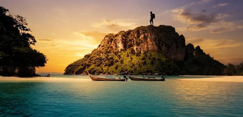 Hiker standing on top of a rocky island above moored longtail boats at sunset