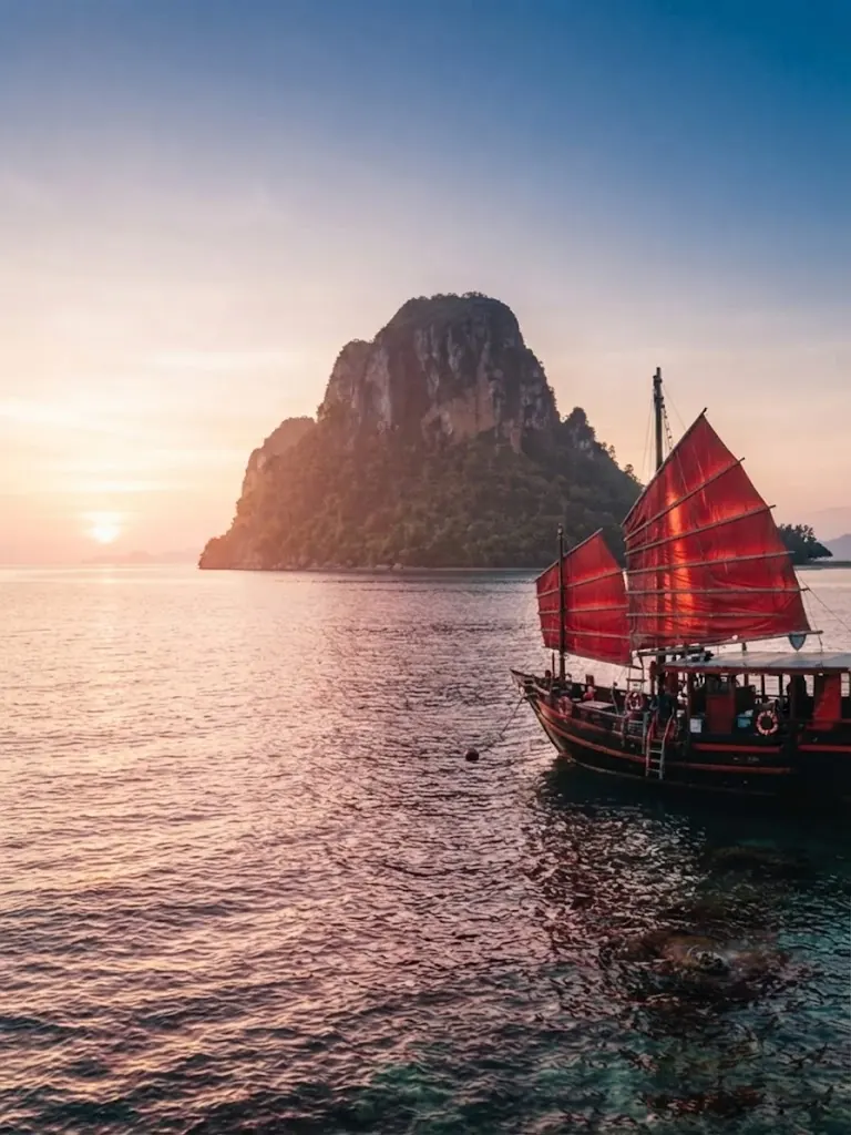 Red-sailed junk boat cruising past a limestone island at sunset