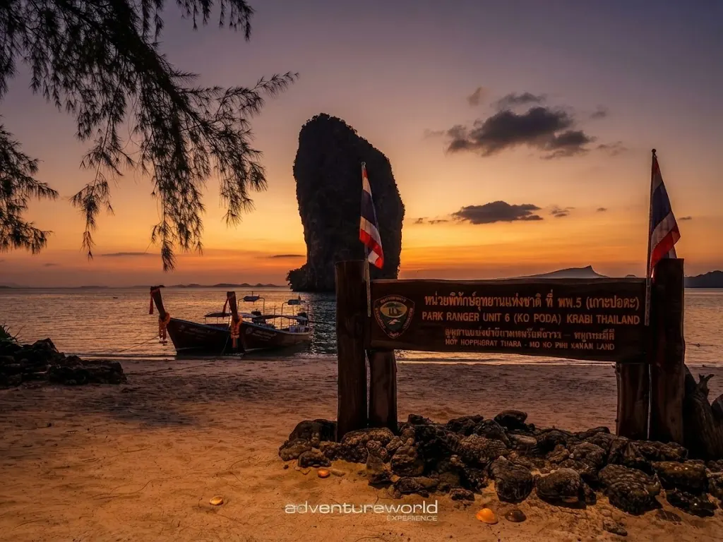 National park ranger station sign for Unit 6, Ko Poda, with Thai flags and a sea stack at sunset