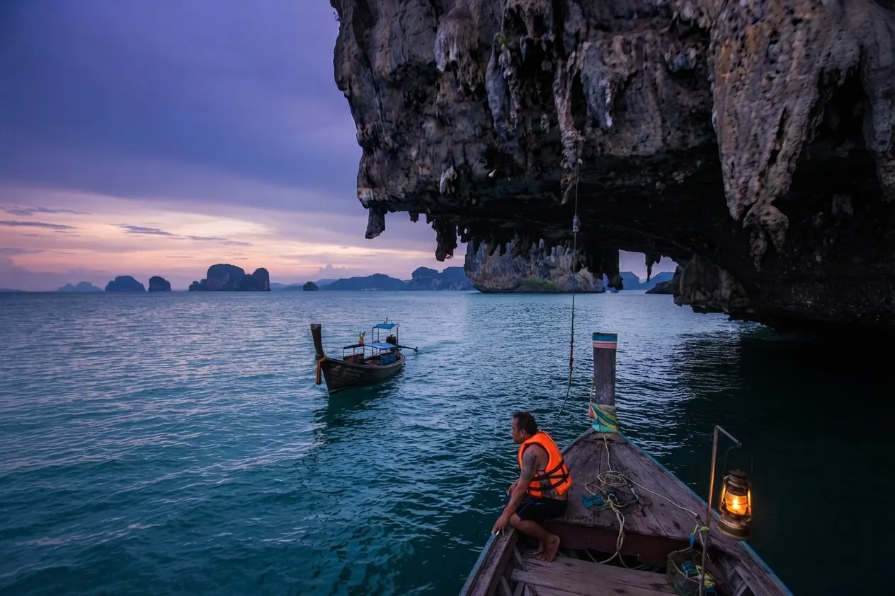 Longtail boatman in a life jacket steering at dusk under a limestone overhang