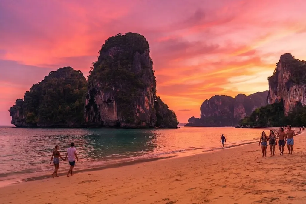 People walking on Phra Nang Beach under a pink sunset sky with limestone islets offshore