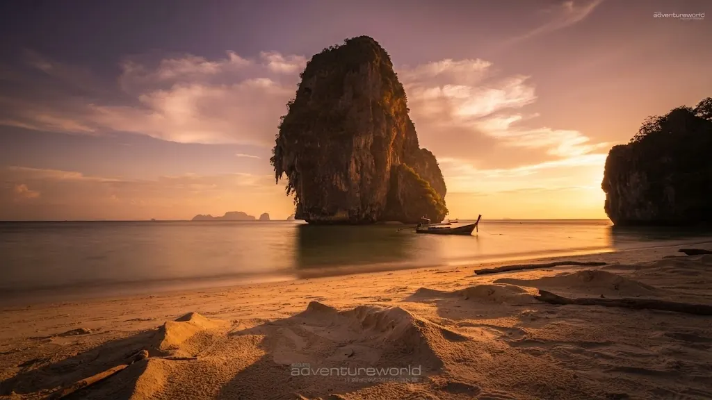 Tall sea stack and a longtail boat off a sandy beach at golden hour near Railay
